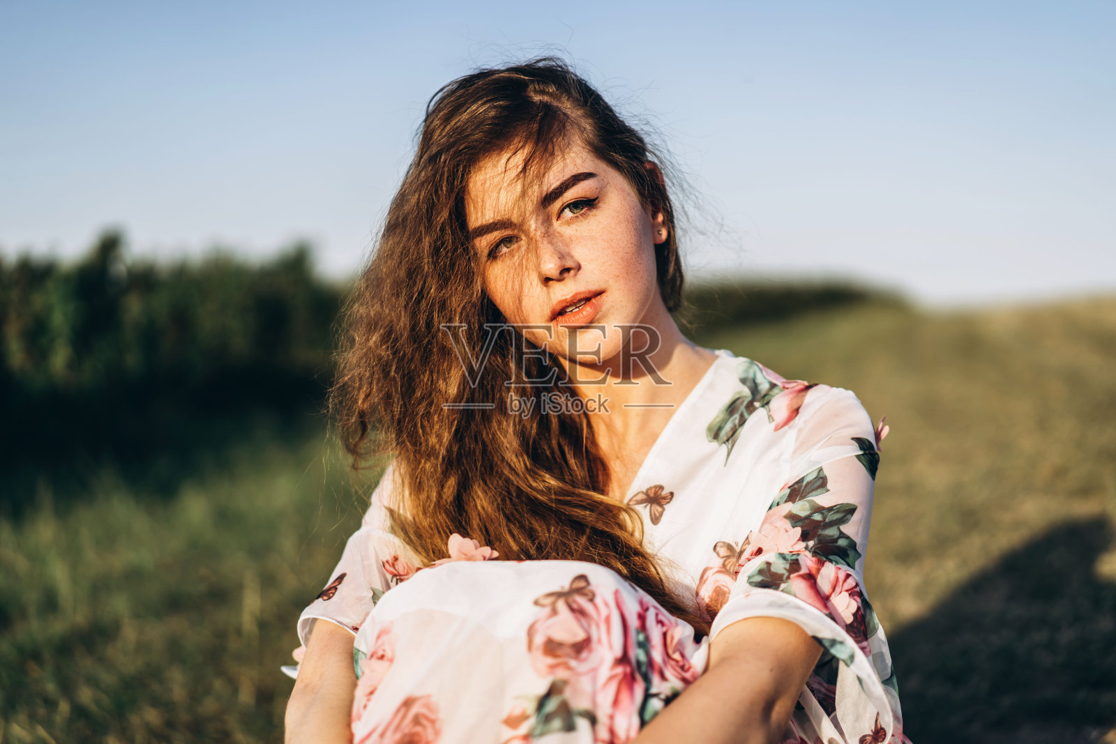 年轻的女孩有着长长的卷发，深色头发，脸上有雀斑，化着妆，有着绿色的眼睛，穿着浅白色的连衣裙在镜头前摆姿势，背景模糊，自然。照片摄影图片