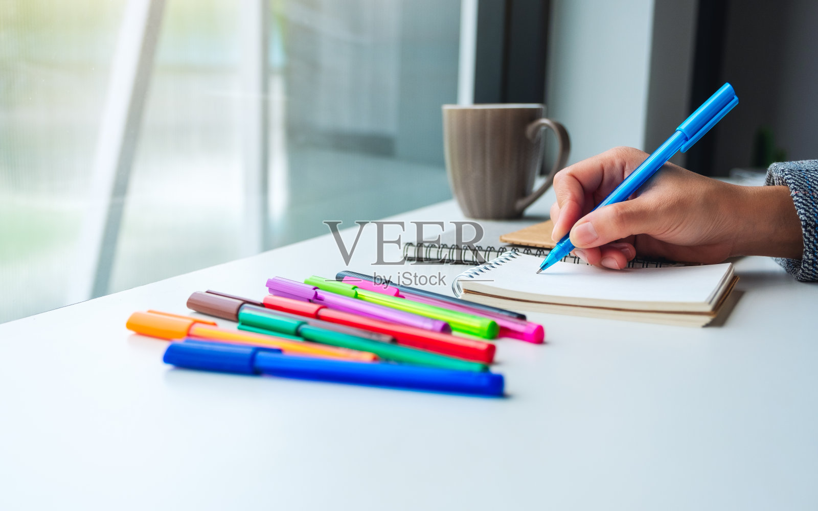 一个女人在一个空白笔记本上写着，桌上放着彩色笔和咖啡杯照片摄影图片