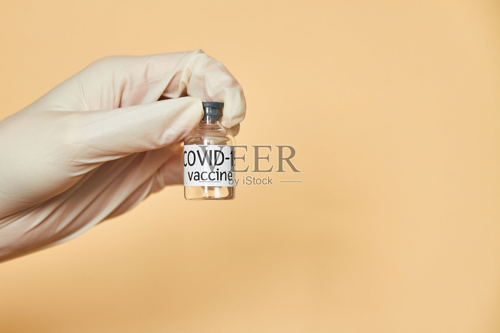 概念上的形象。一只戴着防护手套的手持有Covid-19疫苗。特写镜头,米色背景。医疗保健和医疗理念。照片摄影图片