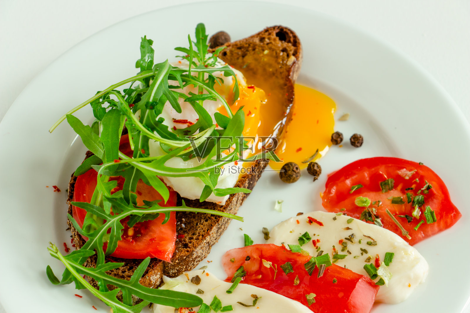美味的荷包蛋放在一片黑麦面包上，配上番茄、马苏里拉奶酪、芝麻菜、香料和黑胡椒，放在一个白色的盘子里照片摄影图片
