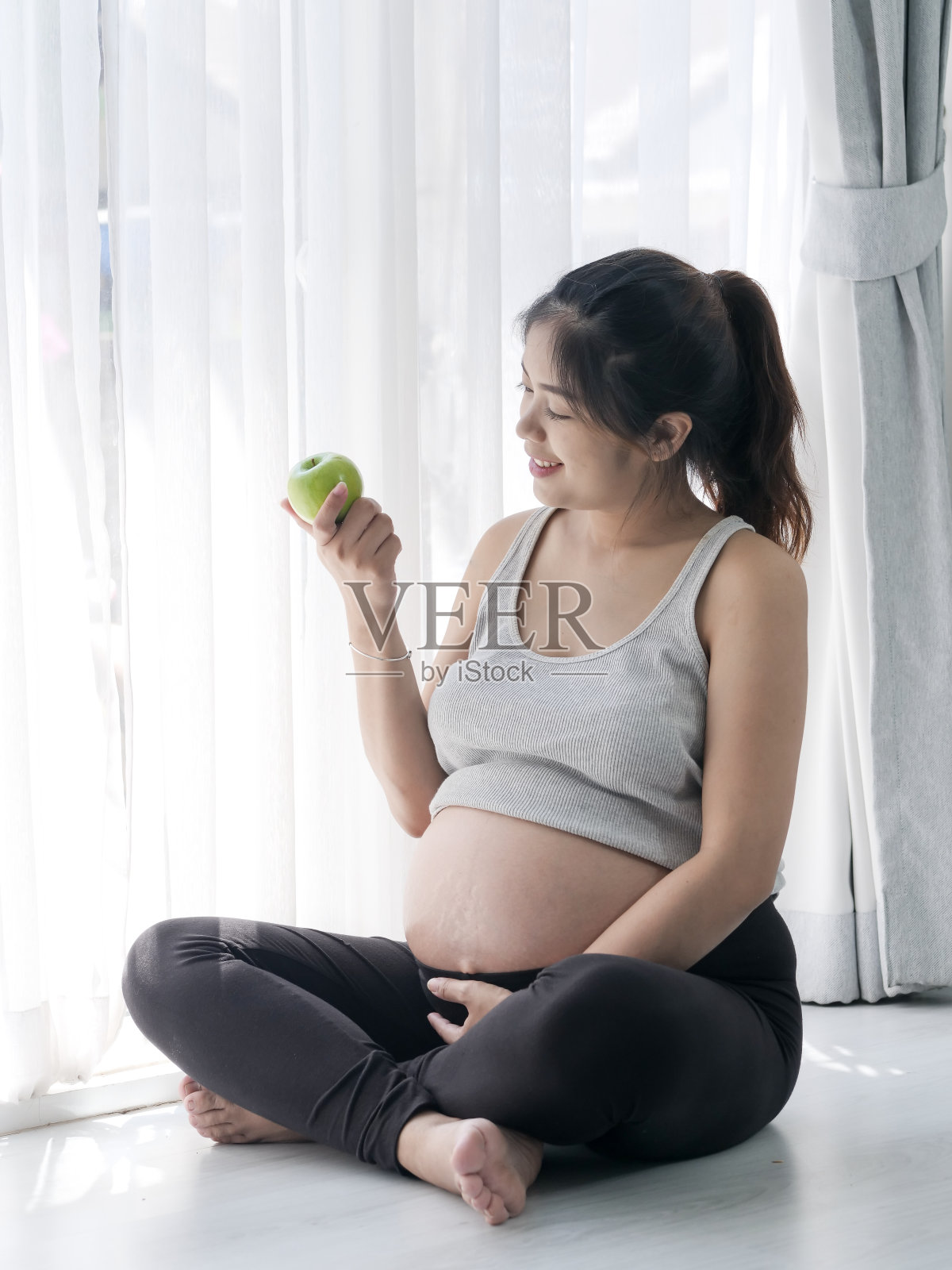 快乐的亚洲女人拿着绿色的苹果在她的手和坐在靠近窗户的地板上，生活理念。照片摄影图片