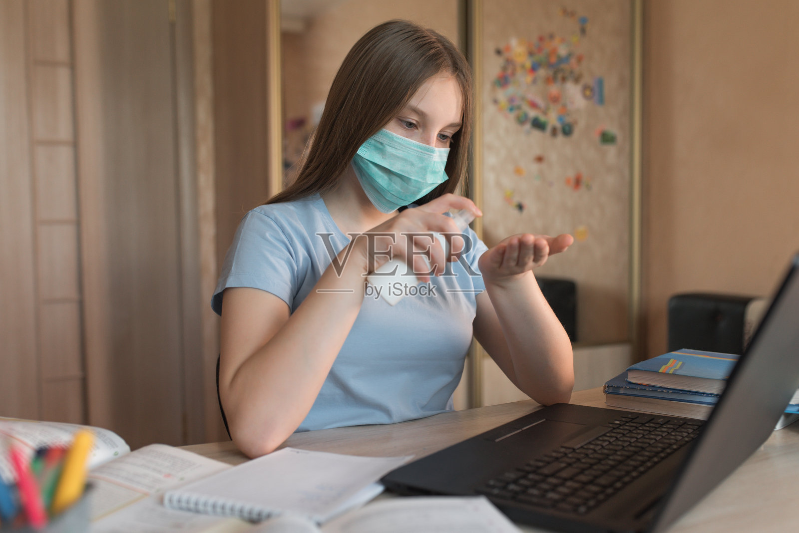 少女戴着医用口罩，用消毒剂喷雾洗手，网上家教视频上网，笔记本电脑上网。隔离期间进行远程教育，待在家里。照片摄影图片