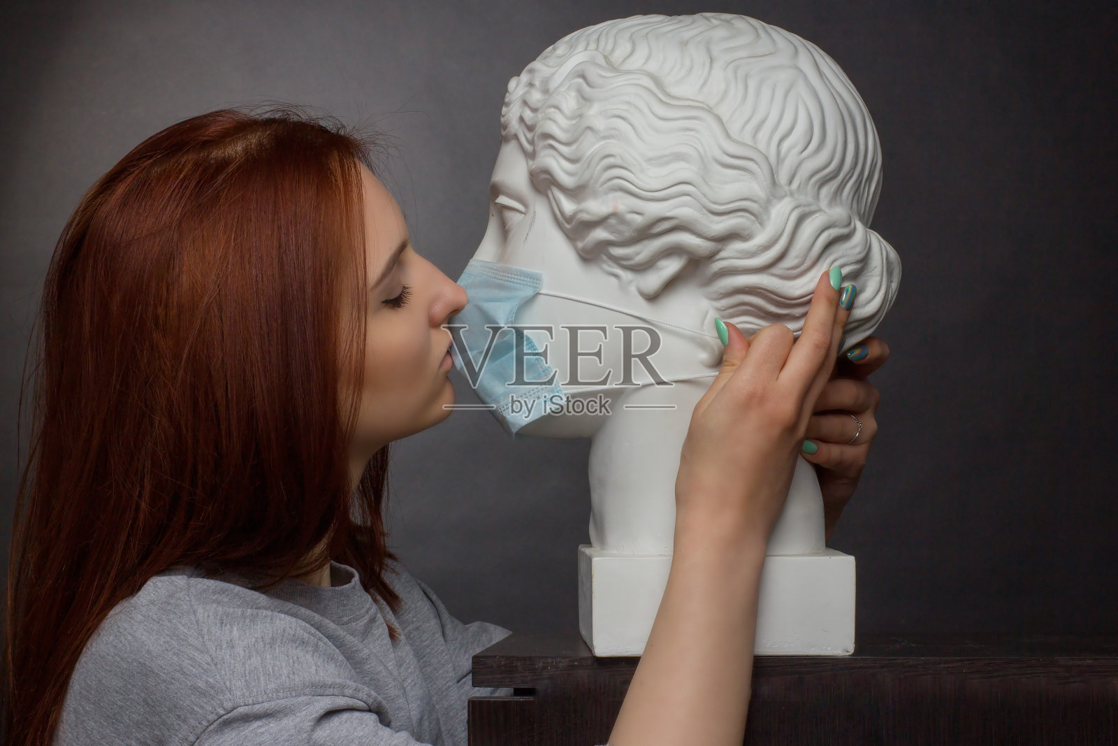 红头发的年轻女孩在一个黑色的背景，灰色的t恤，亲吻与石膏半身像在保护性医疗口罩照片摄影图片