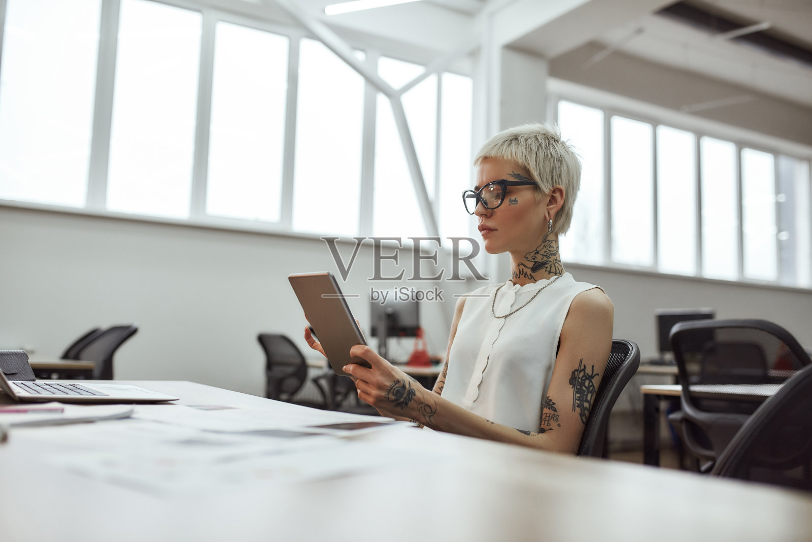 使用数字工具。年轻美丽的纹身商务女性戴着眼镜，坐在她的工作场所，与数字平板电脑照片摄影图片