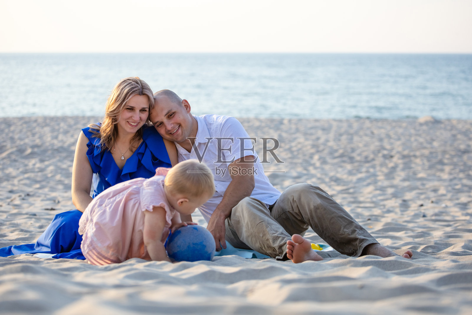 快乐的年轻父母坐在以大海为背景的沙滩上，带着爱看着他们的孩子照片摄影图片