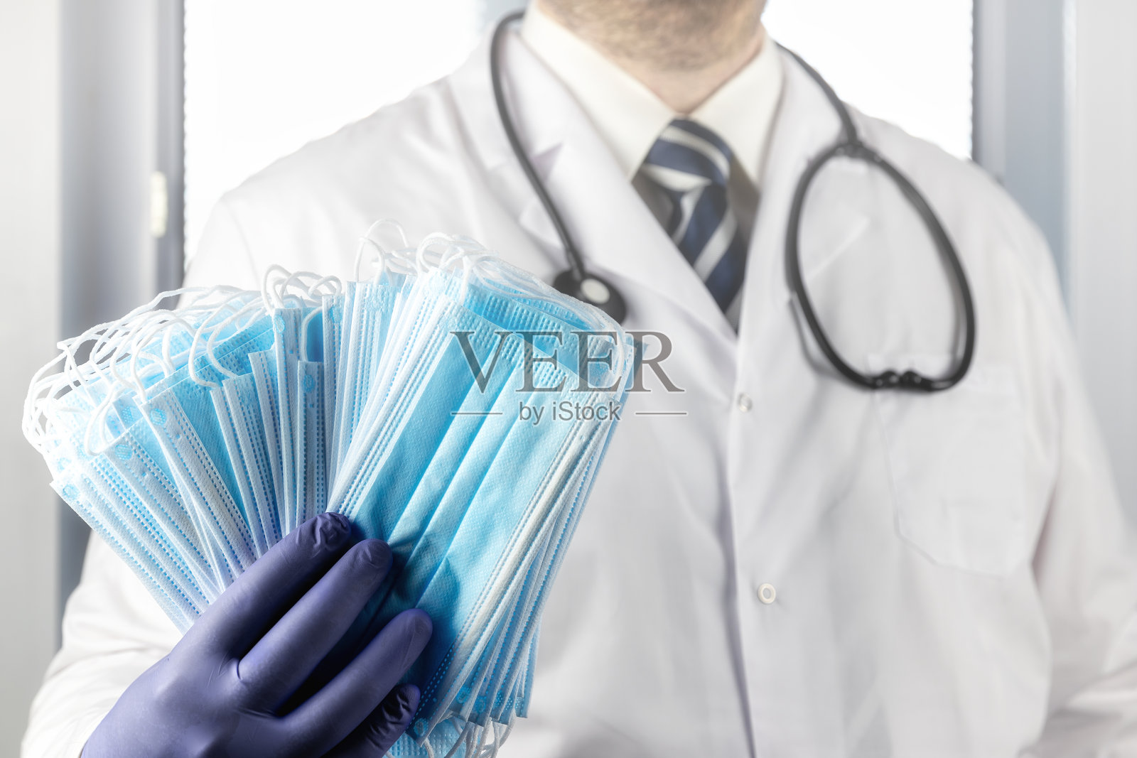 医生身穿白大褂，手持听诊器和手套，手里拿着一叠蓝色医用口罩。医院病房和带百叶窗的窗户照片摄影图片