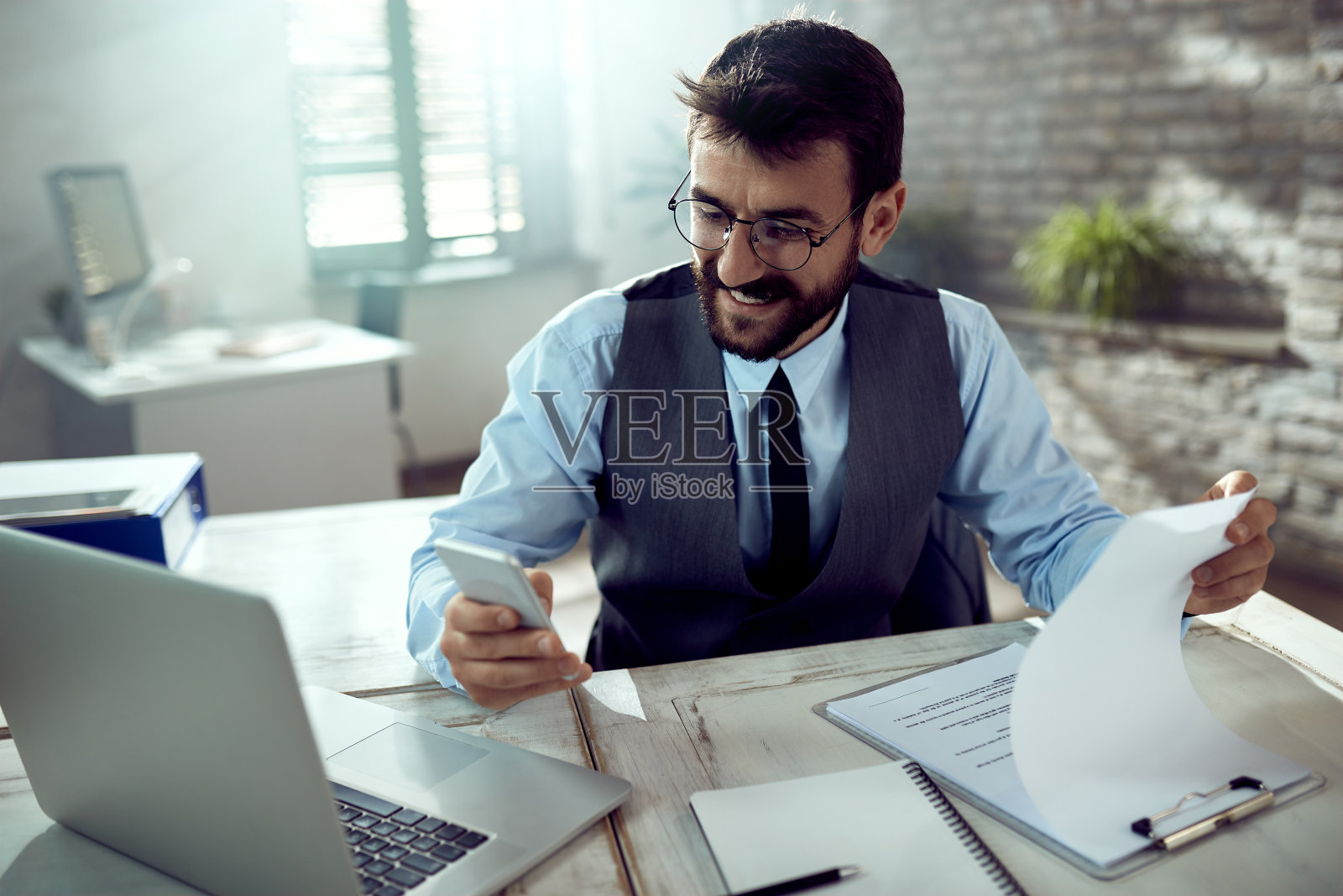 快乐的商人使用手机，同时在办公室做文书工作。照片摄影图片