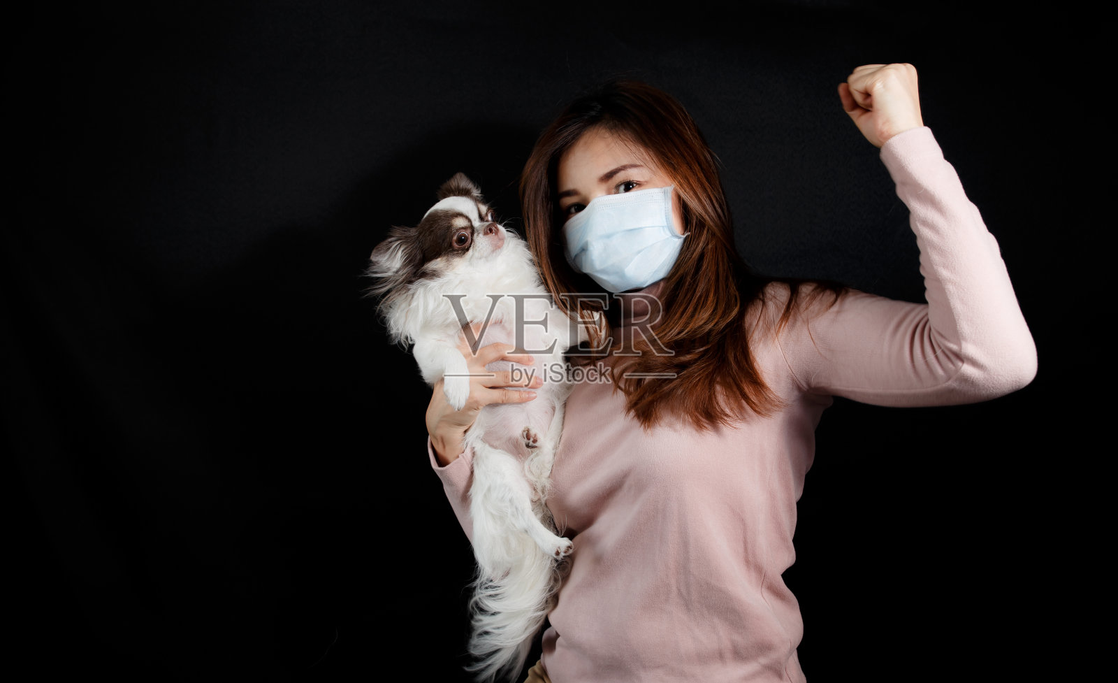 一名妇女戴着口罩，在黑暗的背景下抱着一只狗，以手势表示对抗危机、防雾霾和保护自己免受病毒的信心。冠状病毒大流行。照片摄影图片