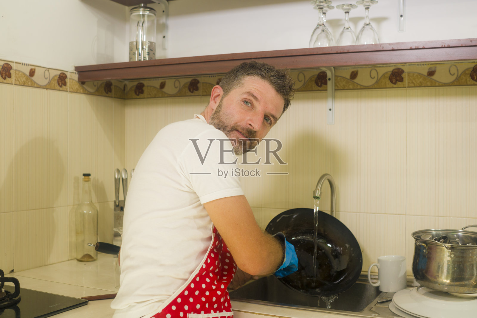 快乐和迷人的男人穿着红围裙微笑在厨房洗盘子和烹饪锅愉快地做家务，享受家庭生活方式照片摄影图片