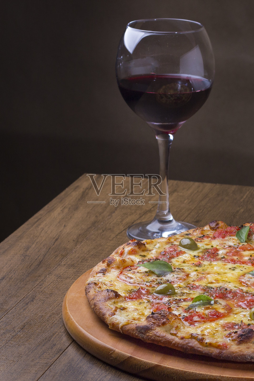 玛格丽特披萨由美味的披萨面团，马苏里拉奶酪，西红柿，马郁兰和绿橄榄制成。放在木板上。Napolitan披萨。红酒盛在玻璃碗里。垂直的照片。照片摄影图片