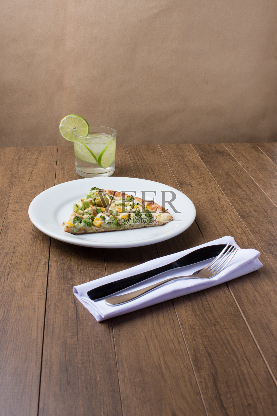 西兰花披萨放在木板上。与马苏里拉奶酪、西兰花、玉米、豌豆和橄榄一起烘烤。美味的披萨放在盘子里，配上一杯柠檬水和刀叉。照片摄影图片