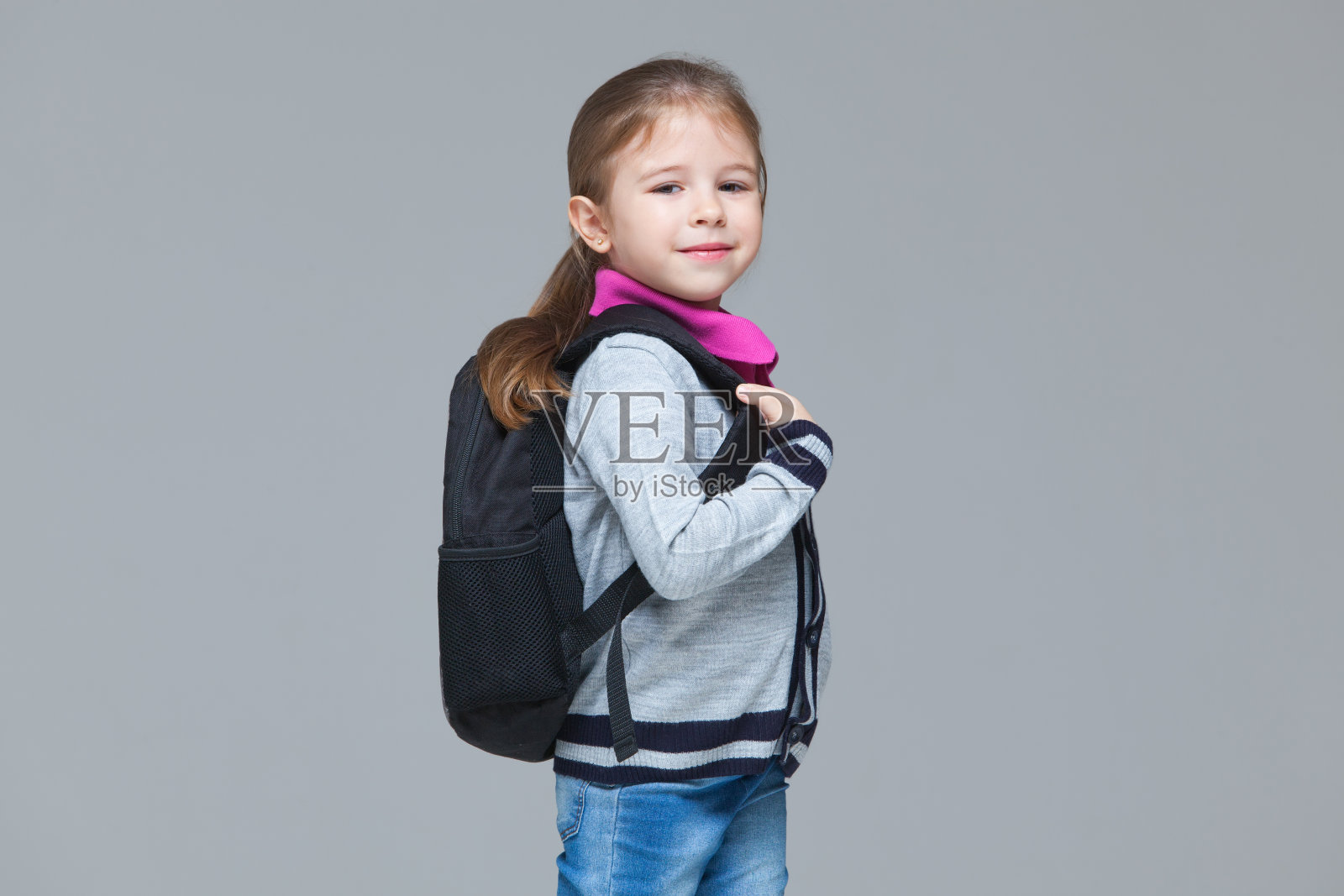 小学女生穿着牛仔裤和校服夹克，背着背包。在灰色背景上孤立照片摄影图片