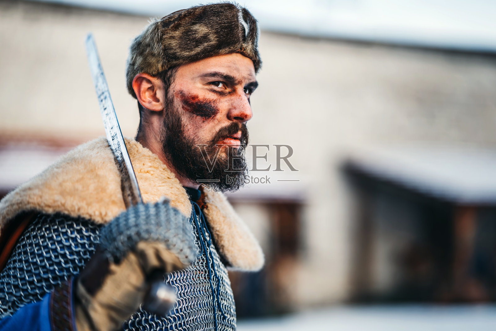 战后身穿盔甲、帽子和披风的年轻战士肖像。一个男人手里拿着武器。在一个木制堡垒的背景在冬天照片摄影图片