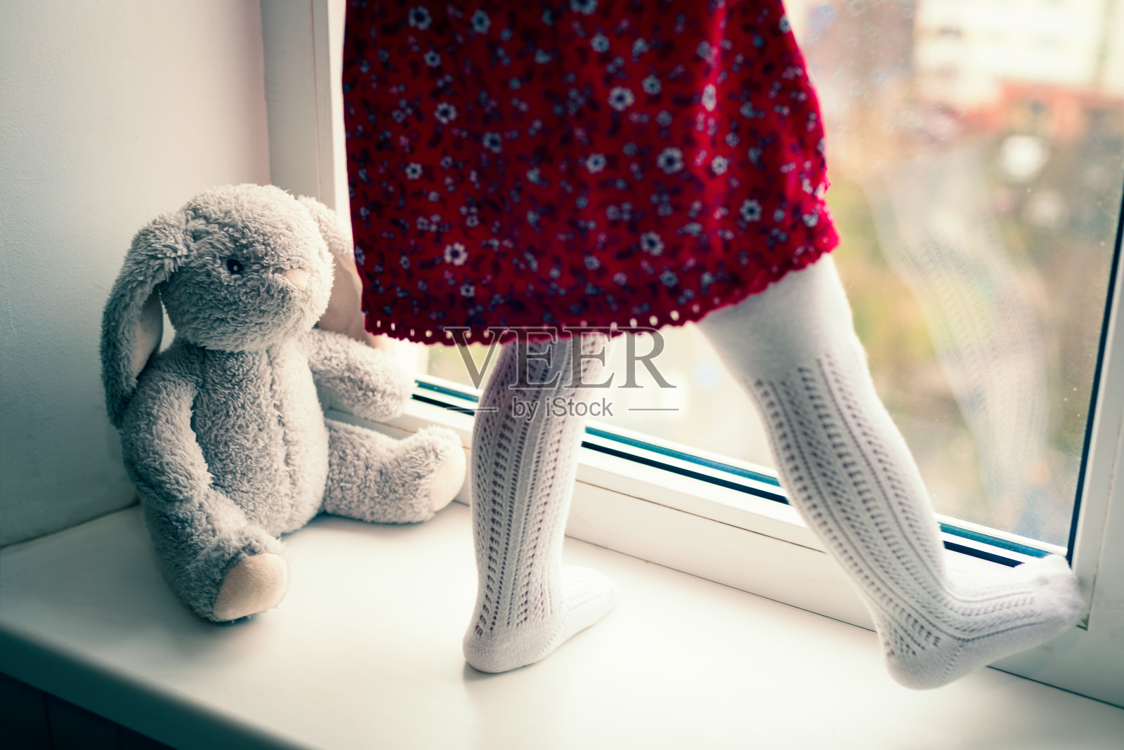 站在窗台上的幼儿和毛绒兔子玩具腿部特写，象征着遗弃、悲伤和孤独。焦点在玩具上。照片摄影图片