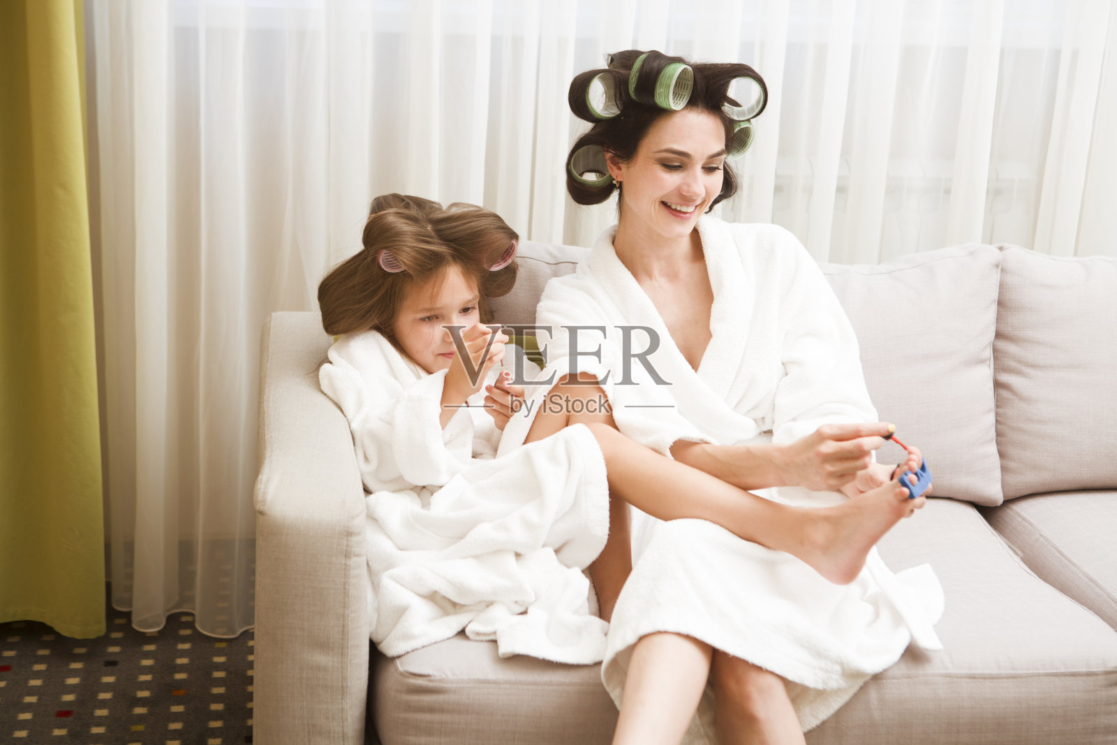 妈妈和女儿穿着白色浴衣，头发上带着卷发器在家做美甲和足疗。他们的头发上有卷发器。快乐的在一起的时间。chelter到位。呆在家里的概念照片摄影图片