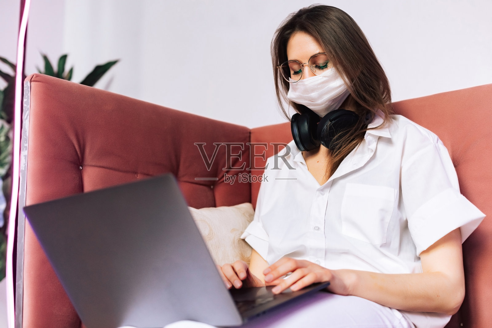 在室内戴着面罩的好女人。她戴一副普通眼镜。概念照片与复制空间。肖像与笔记本电脑和耳机照片摄影图片