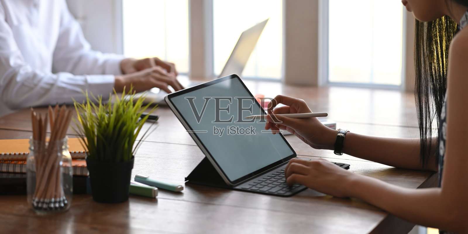 裁剪图像的商务人员工作与白色空白屏幕的平板电脑和笔记本电脑，而坐在一起在木制的办公桌，以舒适的工作室窗户为背景。照片摄影图片