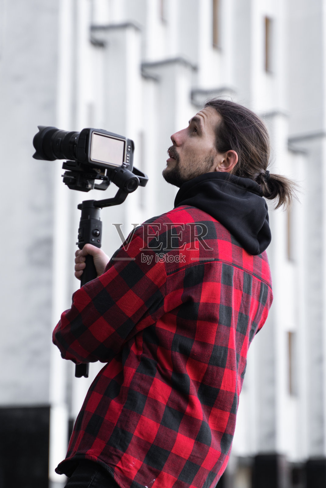 年轻的专业摄像师手持专业摄像机，安装在三轴框架稳定器上。专业设备有助于制作高质量的视频而不摇晃。一名身穿红衬衫的摄影师正在拍摄录像。照片摄影图片