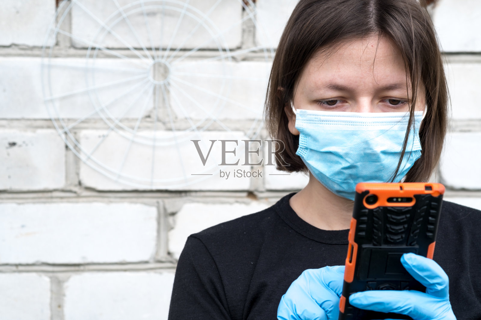 一堵砖墙后面的小女孩戴着防止冠状病毒传播的面罩。妇女在隔离和隔离期间使用电话照片摄影图片