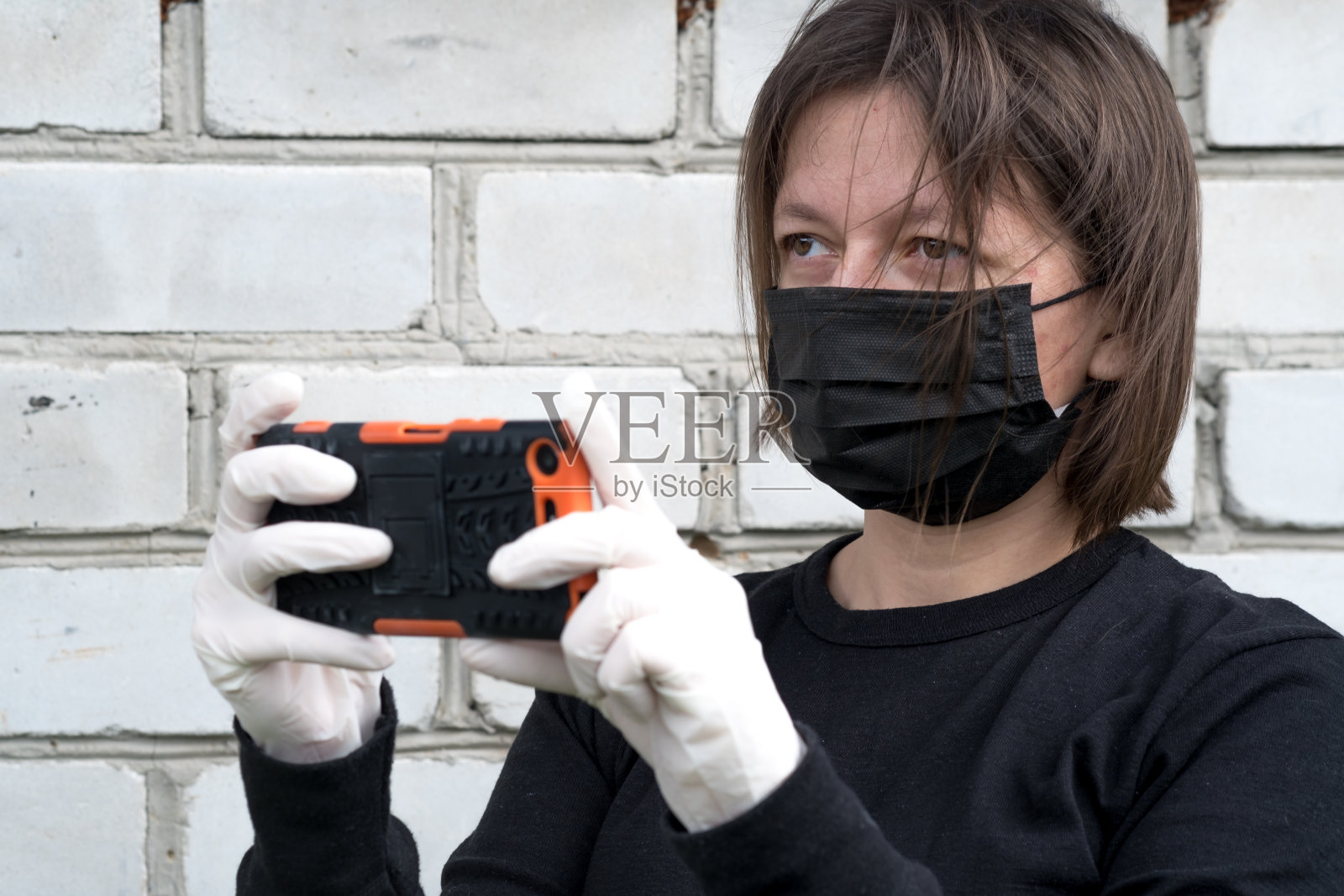 一堵砖墙后面的小女孩戴着防止冠状病毒传播的面罩。妇女在隔离和隔离期间使用电话照片摄影图片