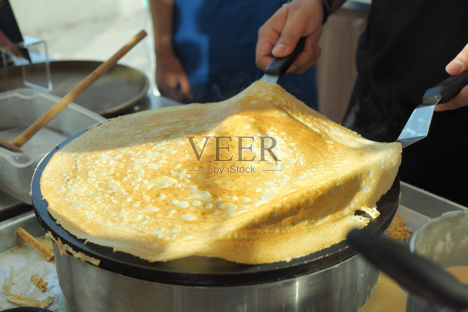 用面粉做可丽饼，倒在热的圆形金属锅上，用金属工具摊开和翻转。照片摄影图片