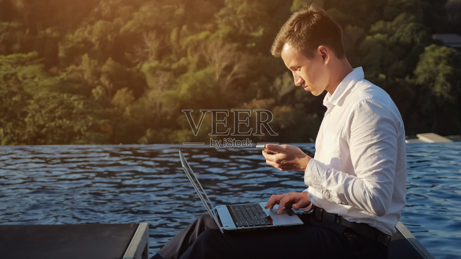 在森林和泳池边，男人在笔记本电脑上工作，看着手机照片摄影图片