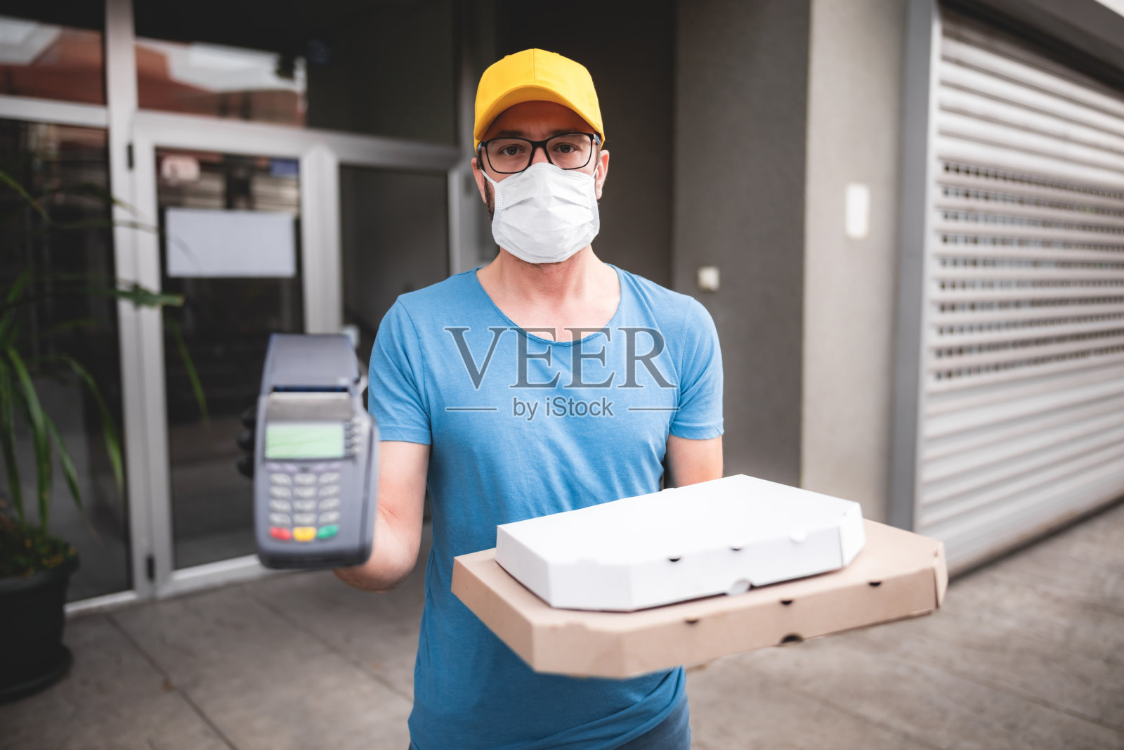 送餐员带防护医用口罩，手持披萨盒和POS无线终端支付卡-病毒和大流行天，食品送至您的家和安全卫生措施。照片摄影图片