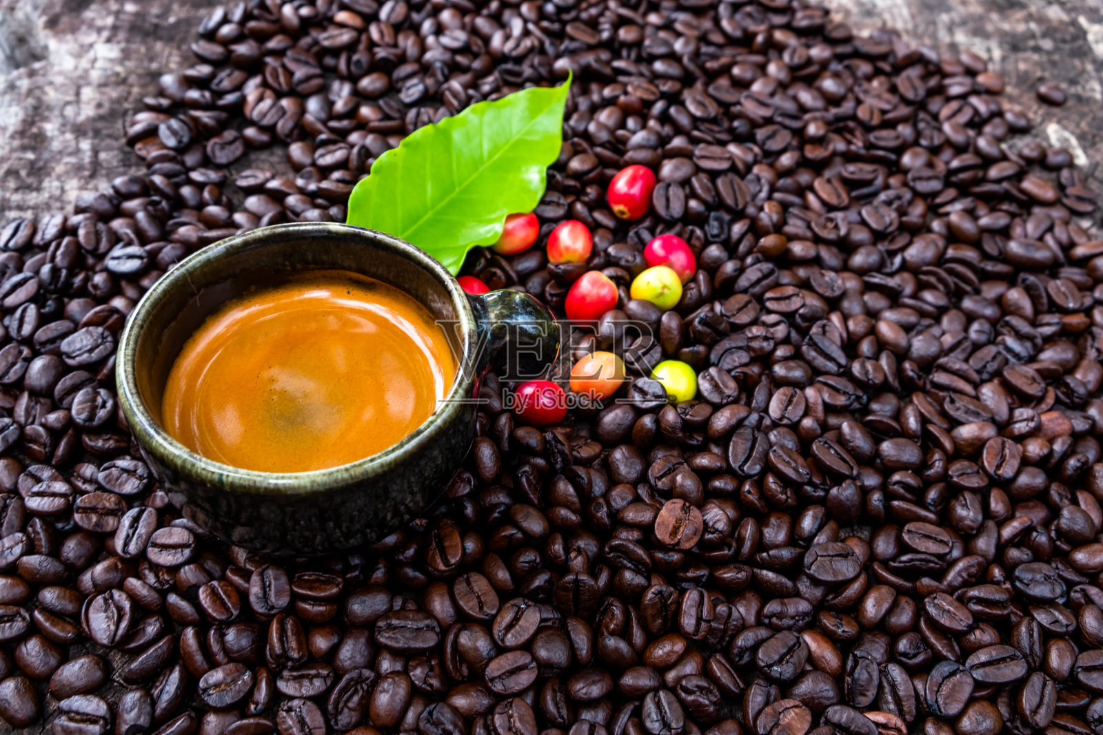 一杯装在咖啡杯里的浓缩咖啡，上面有浅棕色的奶油，一片亮绿色的咖啡叶子，以及散落在深棕色烘焙咖啡豆上的五颜六色的咖啡樱桃。照片摄影图片