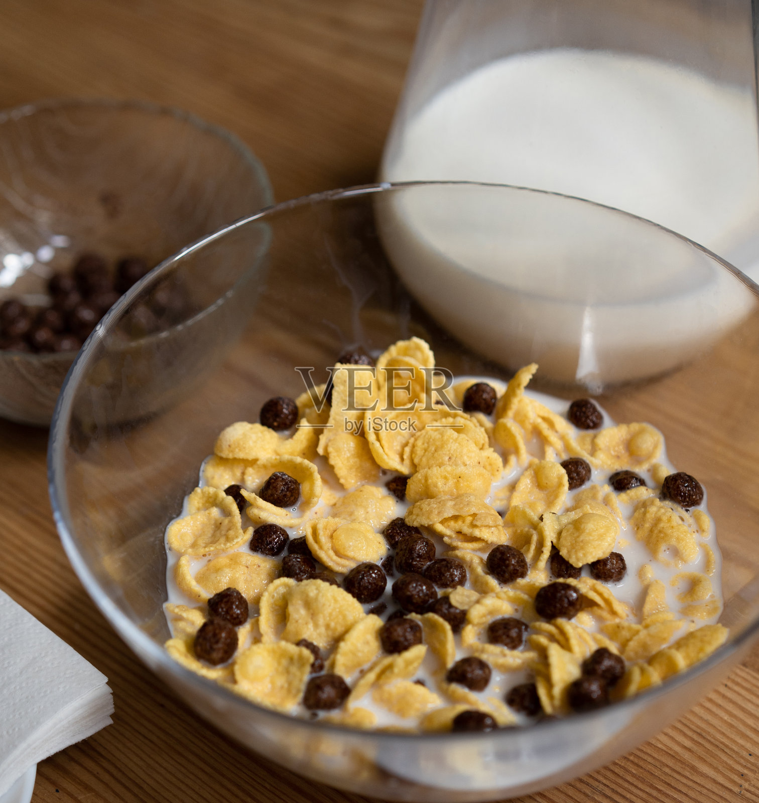 快速的早餐麦片。木桌上的玻璃碗里放着玉米片、巧克力球和牛奶，上面放着一罐牛奶和一碗巧克力球。健康食品的概念照片摄影图片