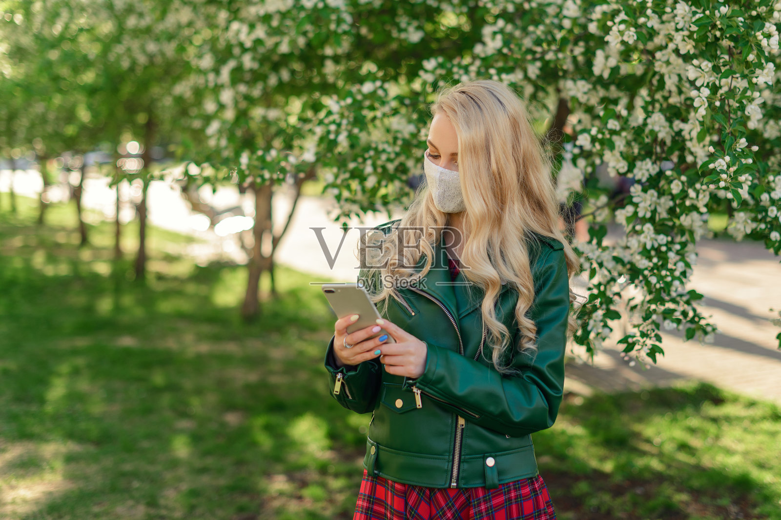 一个金发女孩戴着白色医用口罩，穿着绿色夹克，手里拿着一部白色的智能手机，看着他站在一棵开花的苹果树旁的屏幕照片摄影图片