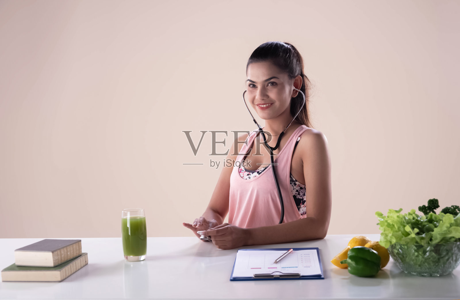 年轻美丽的女士把听诊器绕在她的脖子上，站在果汁杯，书和沙拉碗中间。健康的女人照片摄影图片