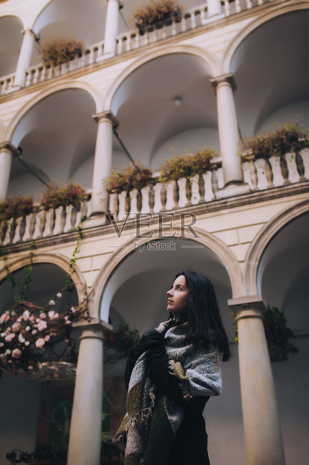一个年轻的白种人黑发女孩，身穿深色秋色外套，裹着宽大的围巾，站在灰色柱廊的背景上，抬起头来。利沃夫，意大利庭院，乌克兰。照片摄影图片