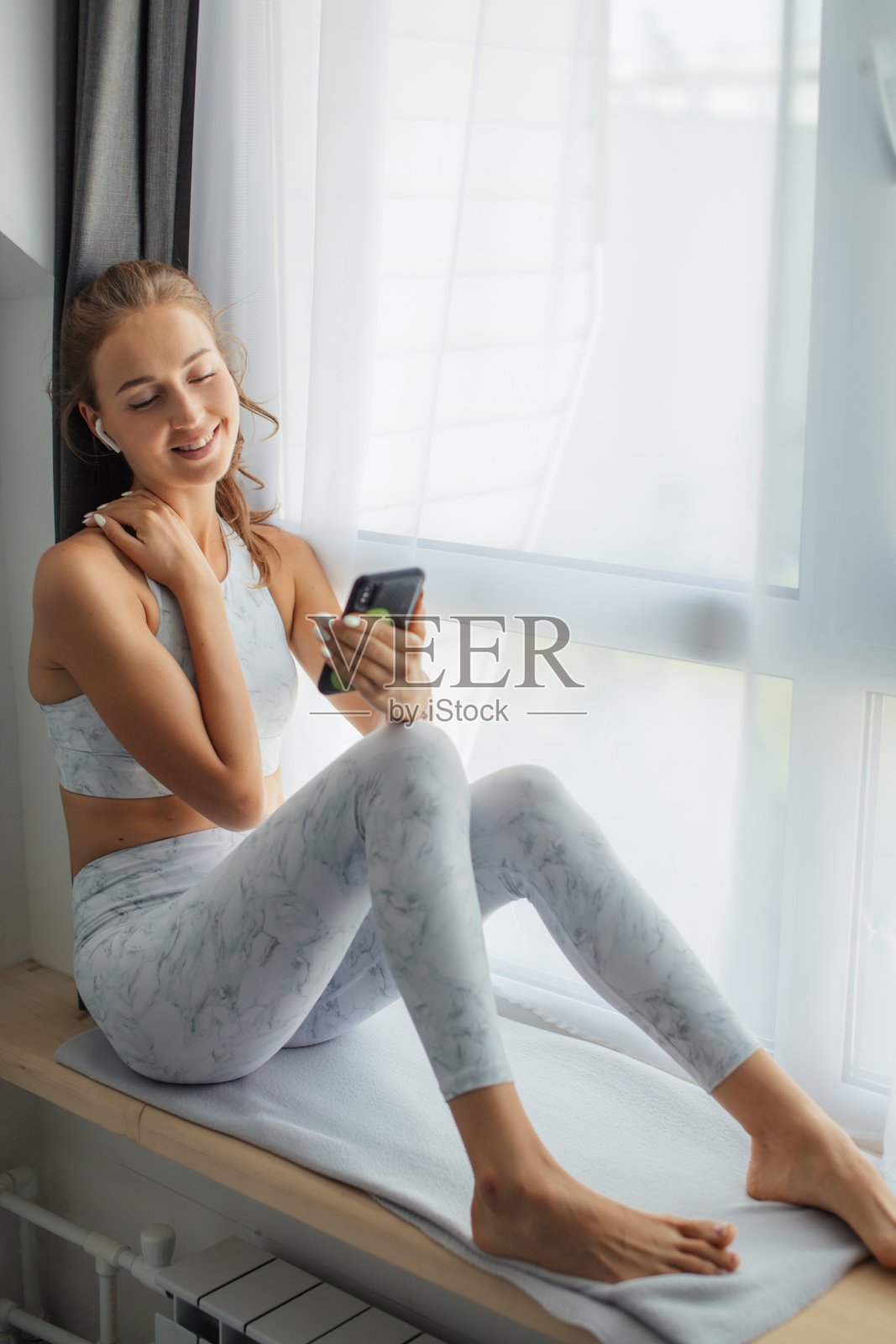 一个女人坐在窗台上用手机和耳机看视频。照片摄影图片