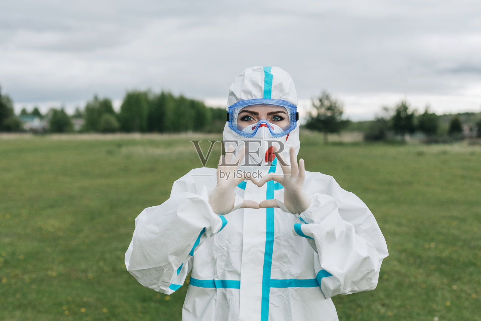 身着防护服的女孩保护自己免受covid-19病毒感染。脸上戴着医用口罩和安全眼镜。手上戴着橡胶手套。持有的心。手语。冠状病毒。隔离。大流行。照片摄影图片