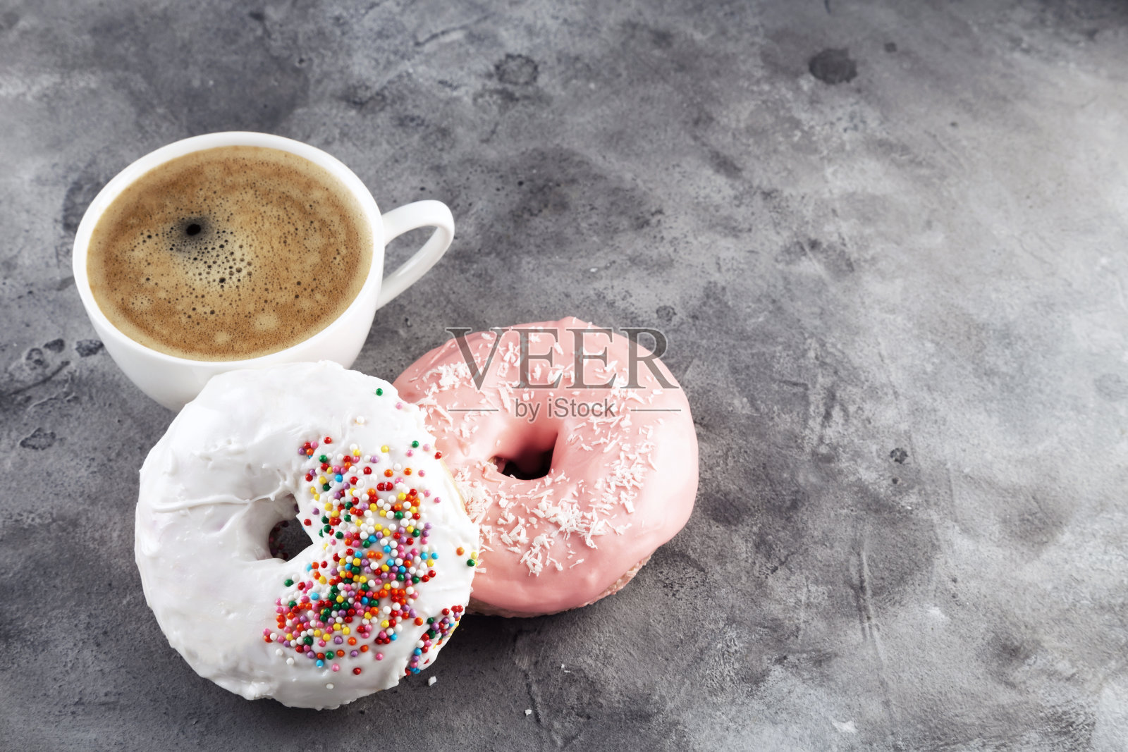 一杯白色的咖啡，两个白色和粉色的糖霜甜甜圈，灰色的背景上点缀着五颜六色的糖糕点。本空间照片摄影图片