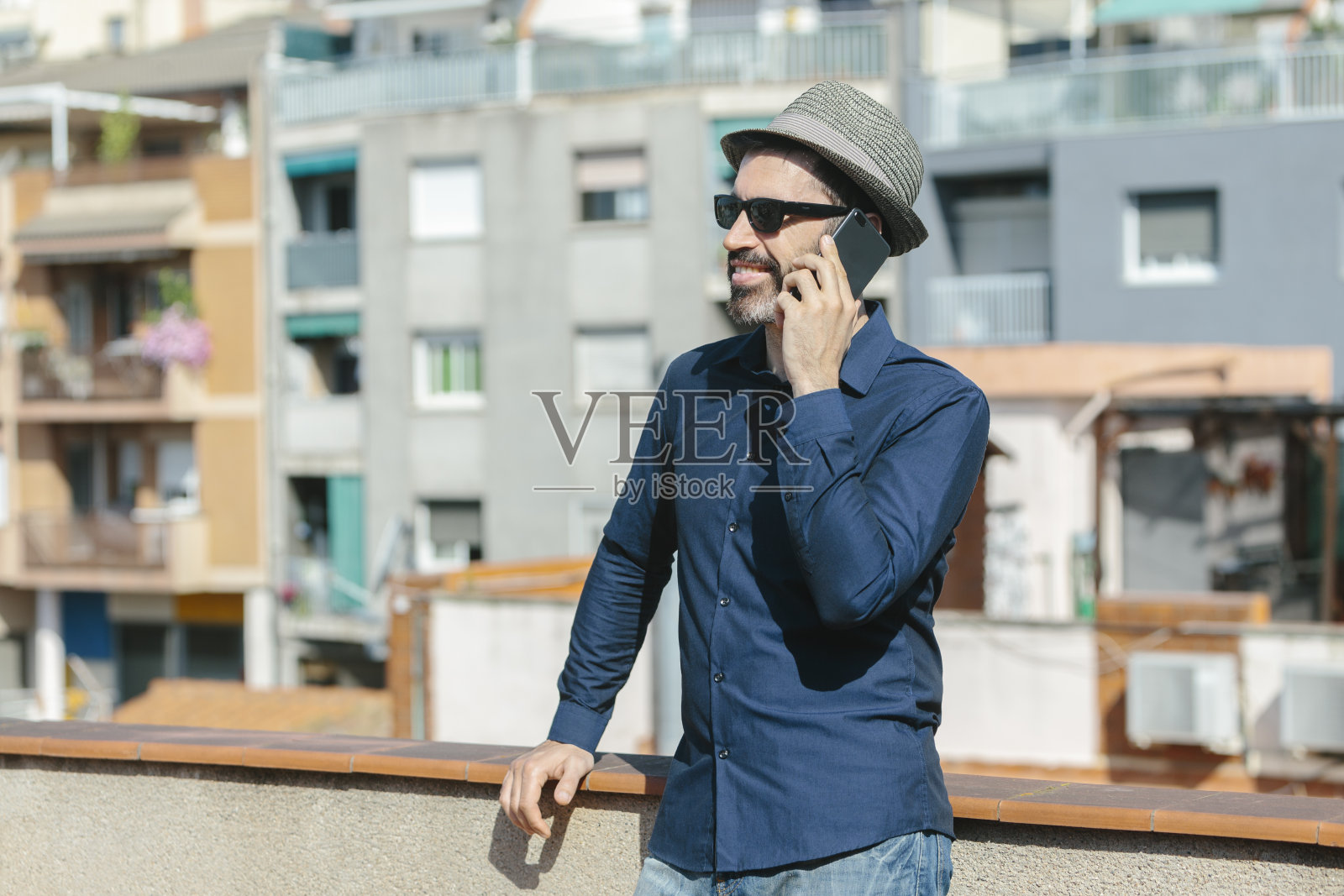 有魅力的中年男子戴着帽子和太阳镜站在阳台上对着智能手机说话。照片摄影图片
