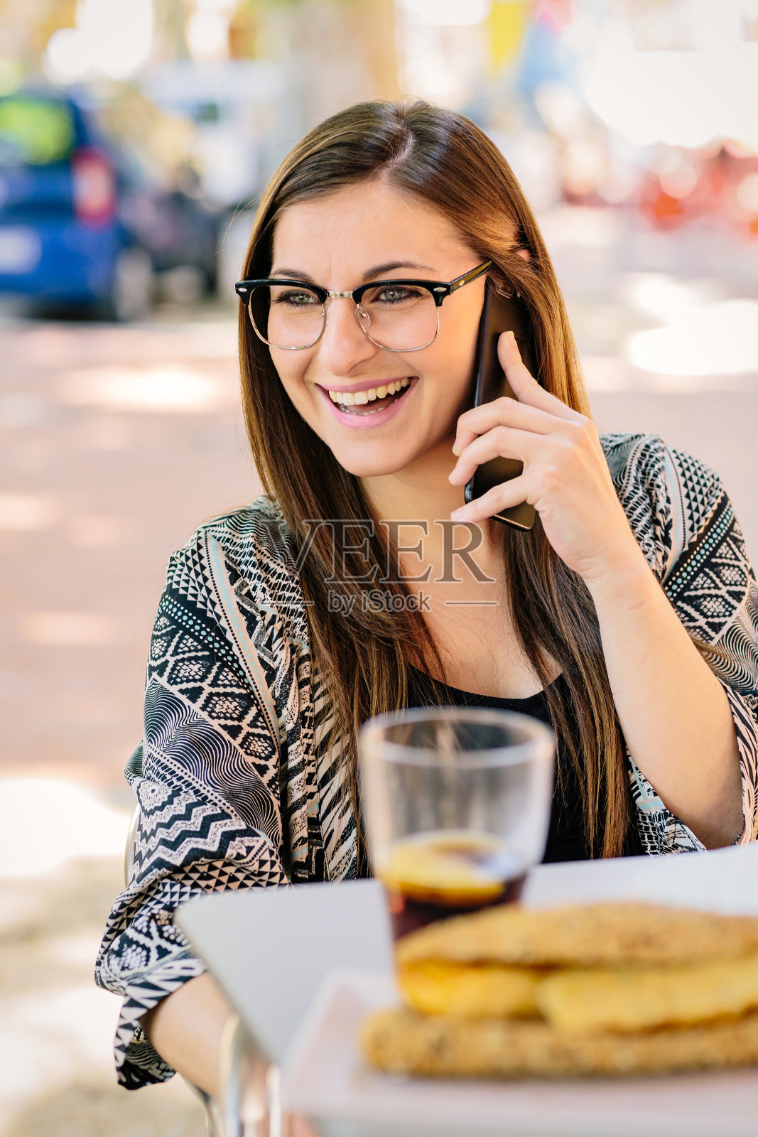 年轻的金发女孩戴着眼镜微笑着与她的朋友讲电话。他正在一家酒吧的露台上喝冷饮。可口可乐照片摄影图片