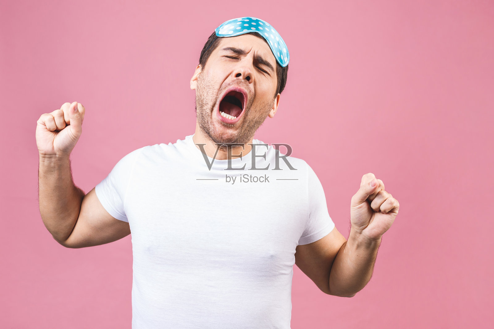 这是一个安静、安静、昏昏欲睡的年轻人的肖像，他戴着蓝色的睡眠面具。孤立的粉红色背景。照片摄影图片