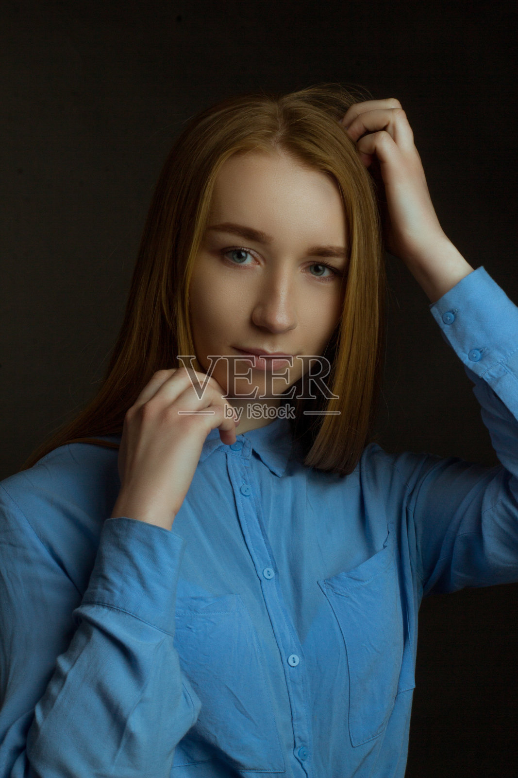 这是一个年轻女孩的肖像，她有着金色的长发，穿着蓝色的衬衫。一个黑暗的形象。照片摄影图片