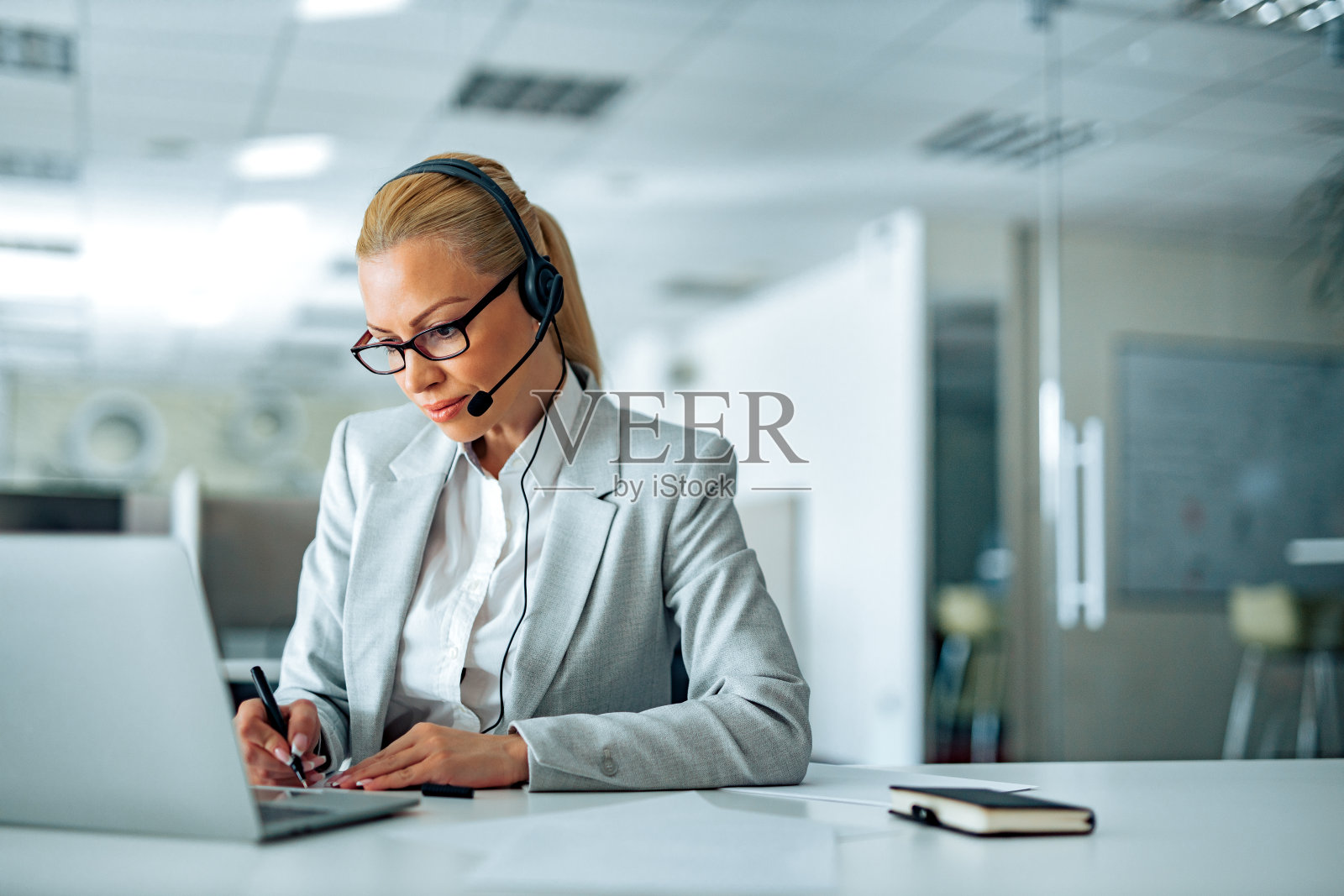 漂亮的金发女商人戴着耳机在笔记本电脑上观看网络研讨会，做笔记。照片摄影图片