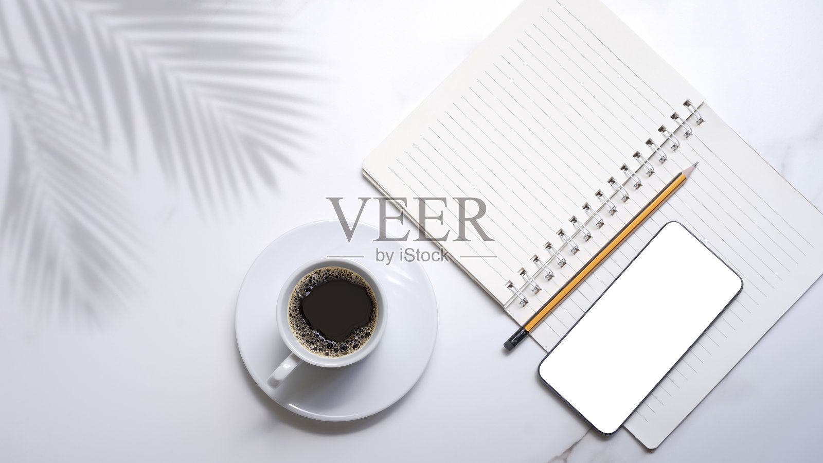 白色陶瓷杯里的黑咖啡，智能手机模型，带螺旋记事本的铅笔，白色大理石桌面上的棕榈叶阴影照片摄影图片