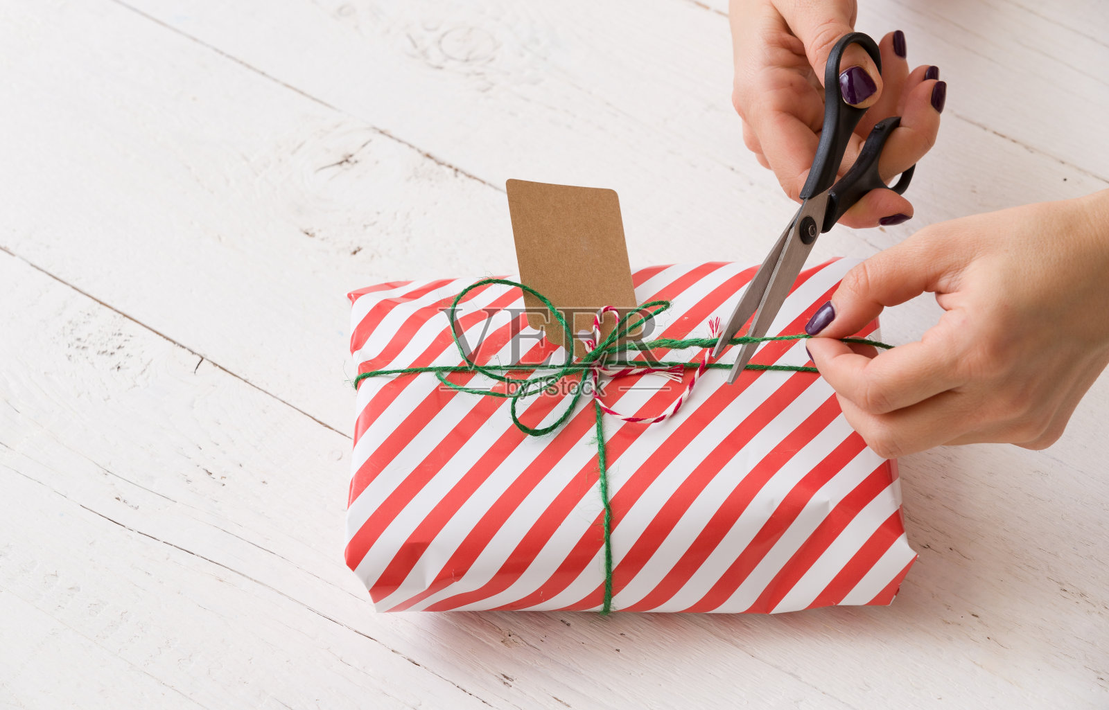 女人的手的特写包装圣诞礼物在条纹纸和装饰它与麻线和标签。女孩在为晚会包装礼物。照片摄影图片