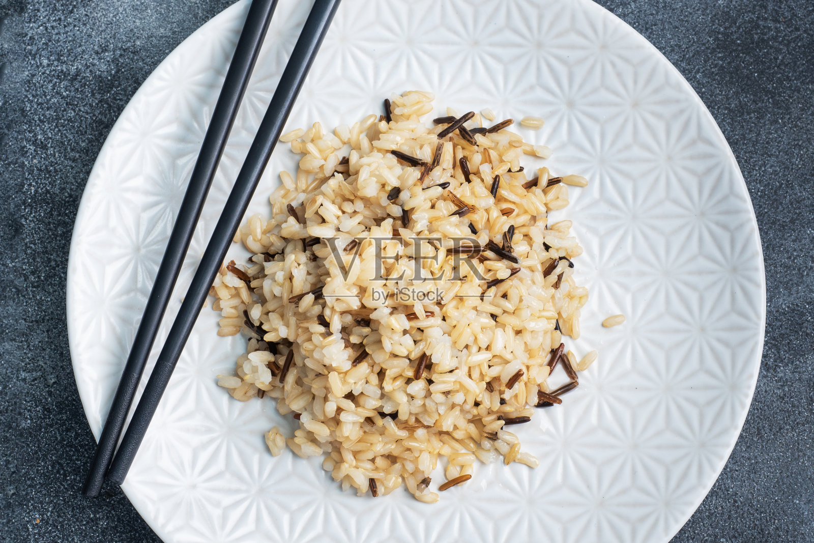 用筷子将糙米和未剥皮的米饭放在盘子里。黑暗的具体背景。副本的空间。照片摄影图片