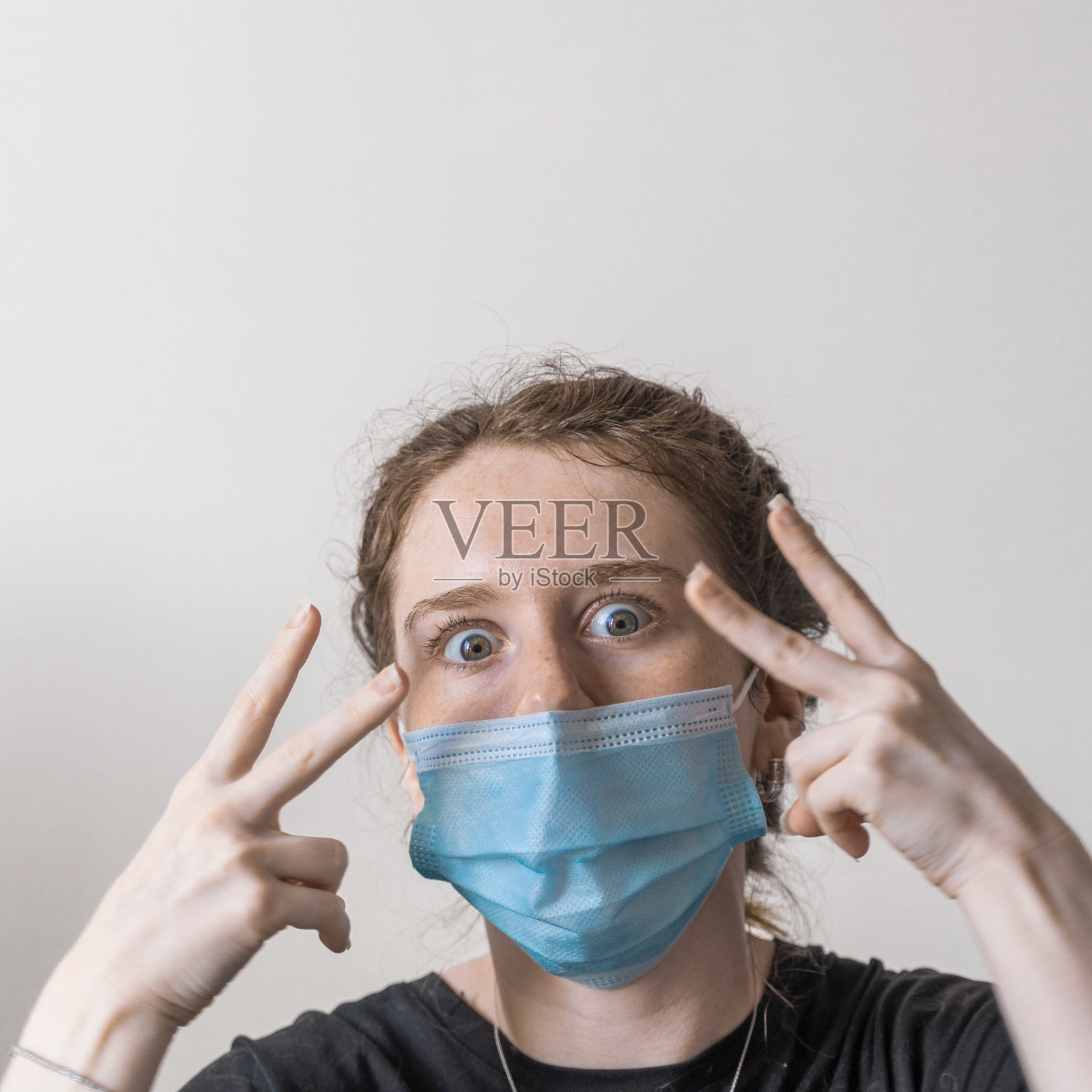 19岁的少女戴着面具，看着镜头，做着滑稽的表情，做出“胜利”和“怎么了”之间的混合手势。照片摄影图片