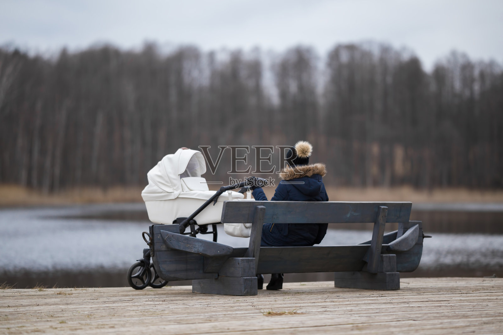 在寒冷的日子里，一个女人坐在木凳上凝视着湖面。白色婴儿推车旁边年轻的母亲。对生活的想法。后视图。宁静的自然氛围。照片摄影图片