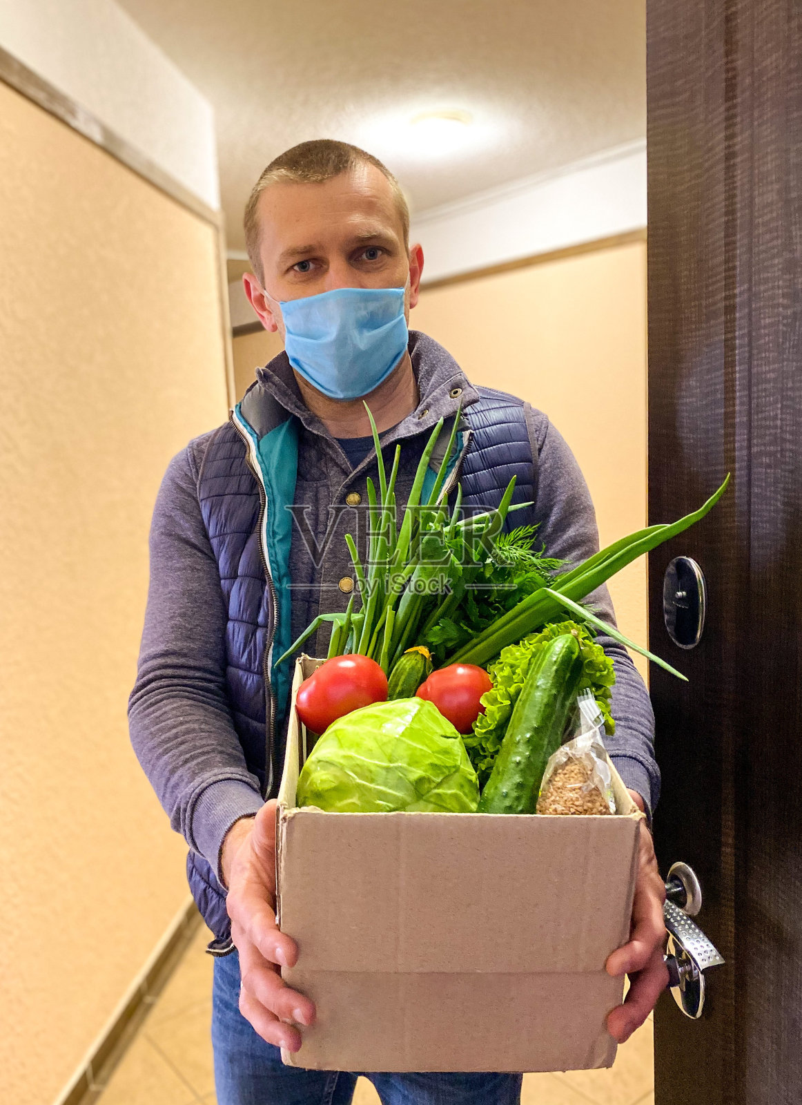 一位皮肤白皙的男子/志愿者戴着医用口罩站在公寓门口，手里拿着一大盒食物。照片摄影图片