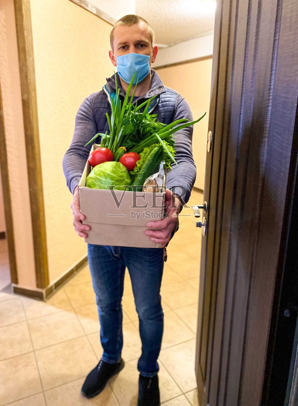 一位皮肤白皙的男子/志愿者戴着医用口罩站在公寓门口，手里拿着一大盒食物。照片摄影图片