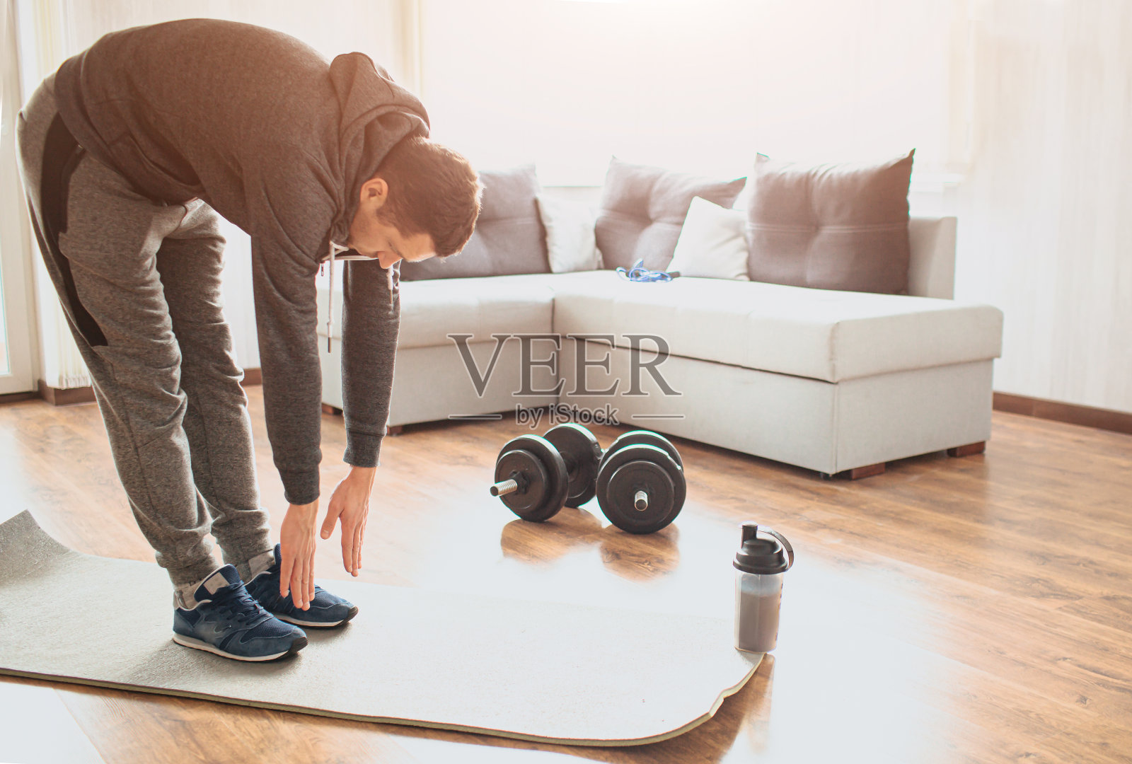 年轻的普通人在家里从事体育运动。站在垫子上，向下伸直脚趾。普通人在运动前暖身。一个人在房间中间锻炼。运动器材在地板上。照片摄影图片