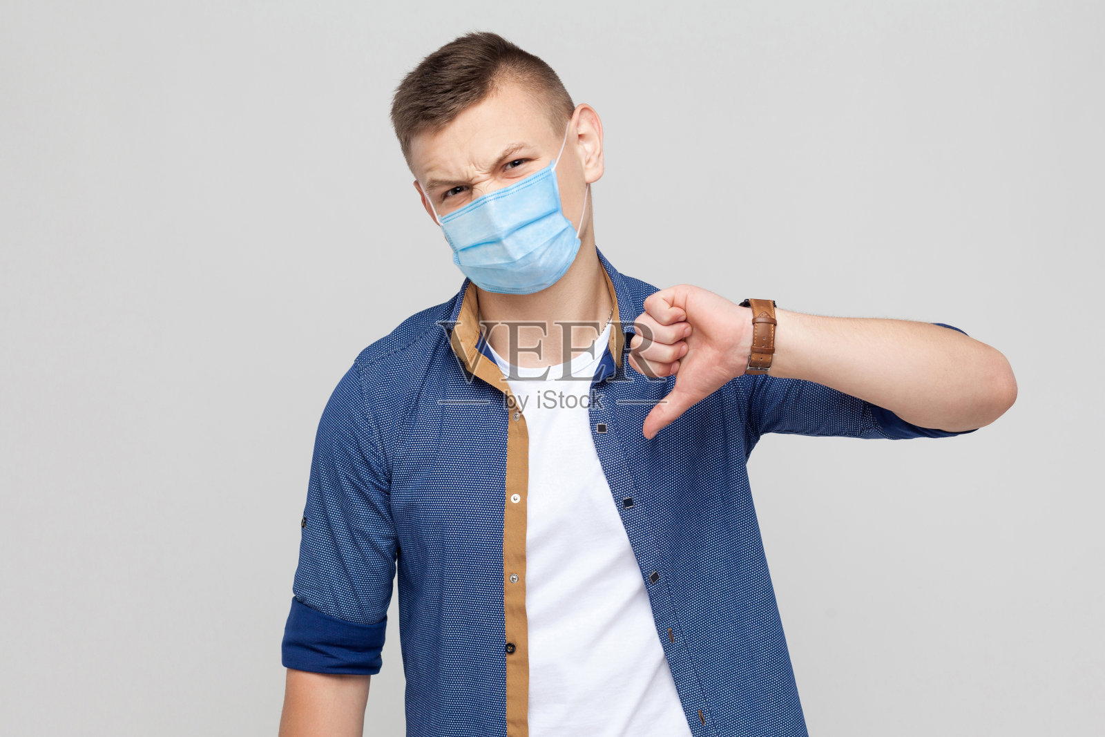 不喜欢。不满意的年轻男子在随意风格与外科医疗口罩站拇指向下，看着相机与不安的脸。照片摄影图片