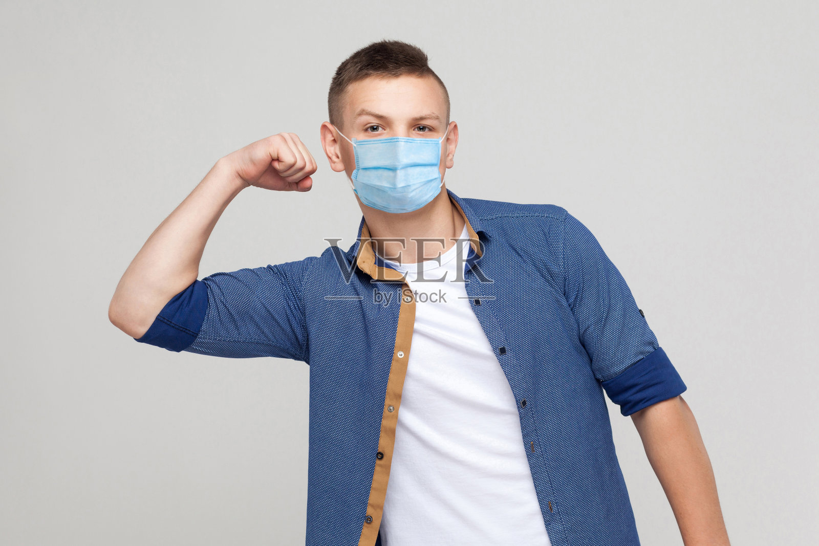 我是坚强的。肖像傲慢的年轻人在随意风格与外科医疗口罩站举起手臂，看着相机与骄傲的脸。照片摄影图片